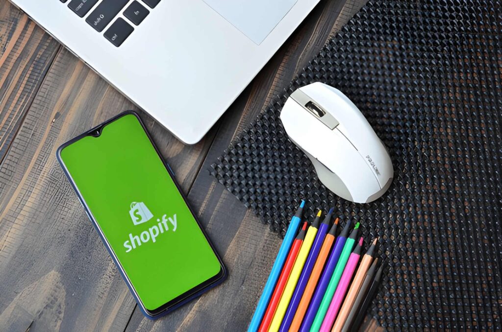Smartphone displaying Shopify app logo next to laptop, mouse, and colored pencils on wooden desk
